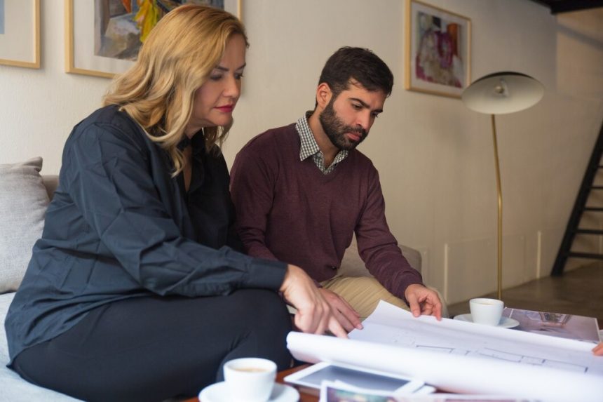 A man and woman sit on a couch, examining a blueprint together with focused expressions